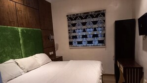 Una habitación con una cama, cabecera verde, armario de madera, una mesita, televisión y persiana con diseño.