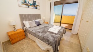 Quarto com cama, mesas de cabeceira, um quadro e vista para o mar através de uma porta deslizante.