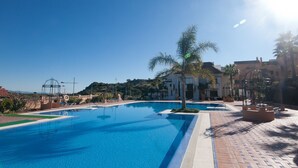 Uma grande piscina ao ar livre com água azul cristalina, rodeada por um calçadão e palmeiras.
