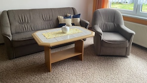 A living room with a grey sofa, a matching armchair, and a wooden coffee table with a placemat. A framed landscape painting is on the wall. - Cozy vacation apartment in the hinterland, ideal for cyclists  (Garz)
