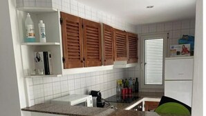 Una cocina con paredes de azulejos blancos, armarios de madera, encimera de granito y una ventana con persianas.