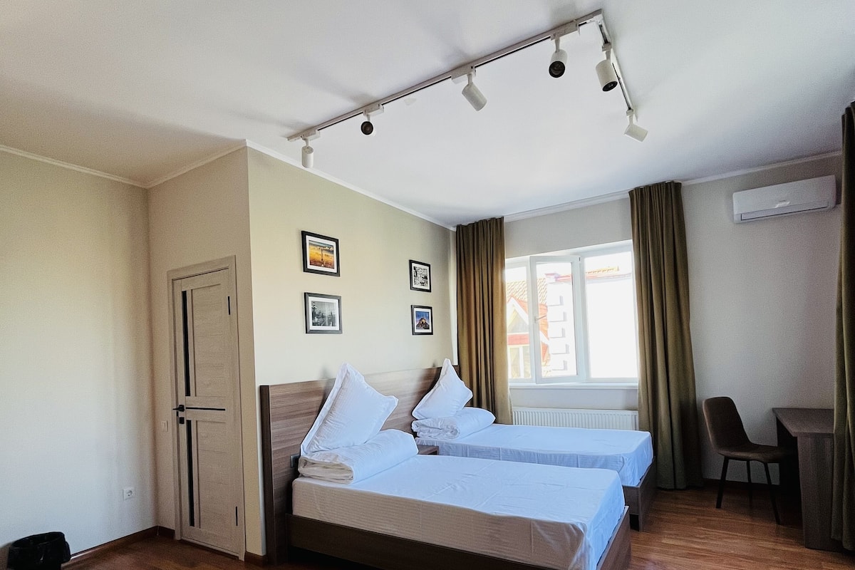 Une chambre d’hôtel avec deux lits, un bureau, une chaise, une fenêtre avec des rideaux et un luminaire au plafond.