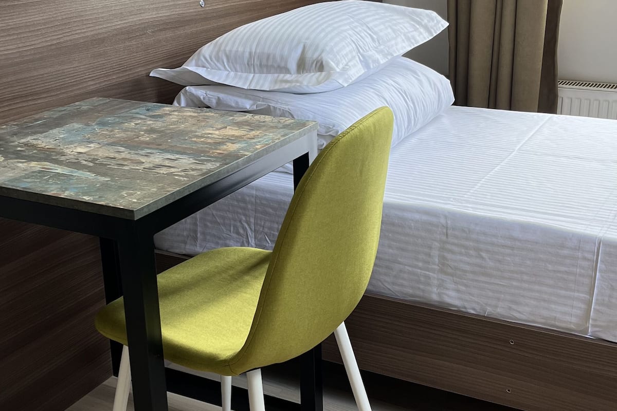 Une chambre avec une tête de lit en bois, un lit recouvert de linge de lit blanc, une chaise verte et une table dont le plateau est orné d’un motif.