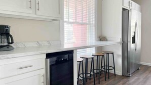 A modern kitchen with white cabinetry, a stainless steel refrigerator, a coffee maker, and a window with blinds.
