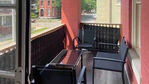 A balcony with two chairs and a table, a red brick wall, and a view of a street with buildings.