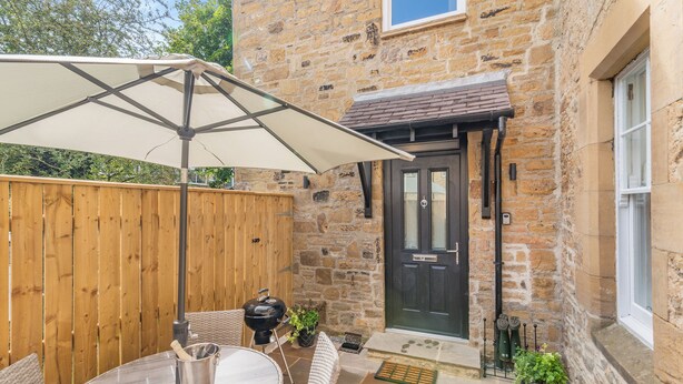 Un bâtiment en pierre avec une porte noire, une clôture en bois, une terrasse avec des meubles en osier et une table sur laquelle se trouve un seau en métal.