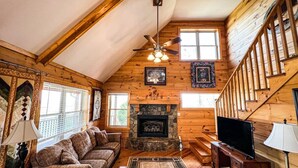 L’intérieur chaleureux d’un chalet, avec un revêtement mural en bois, une cheminée en pierre, un ventilateur de plafond et un escalier.