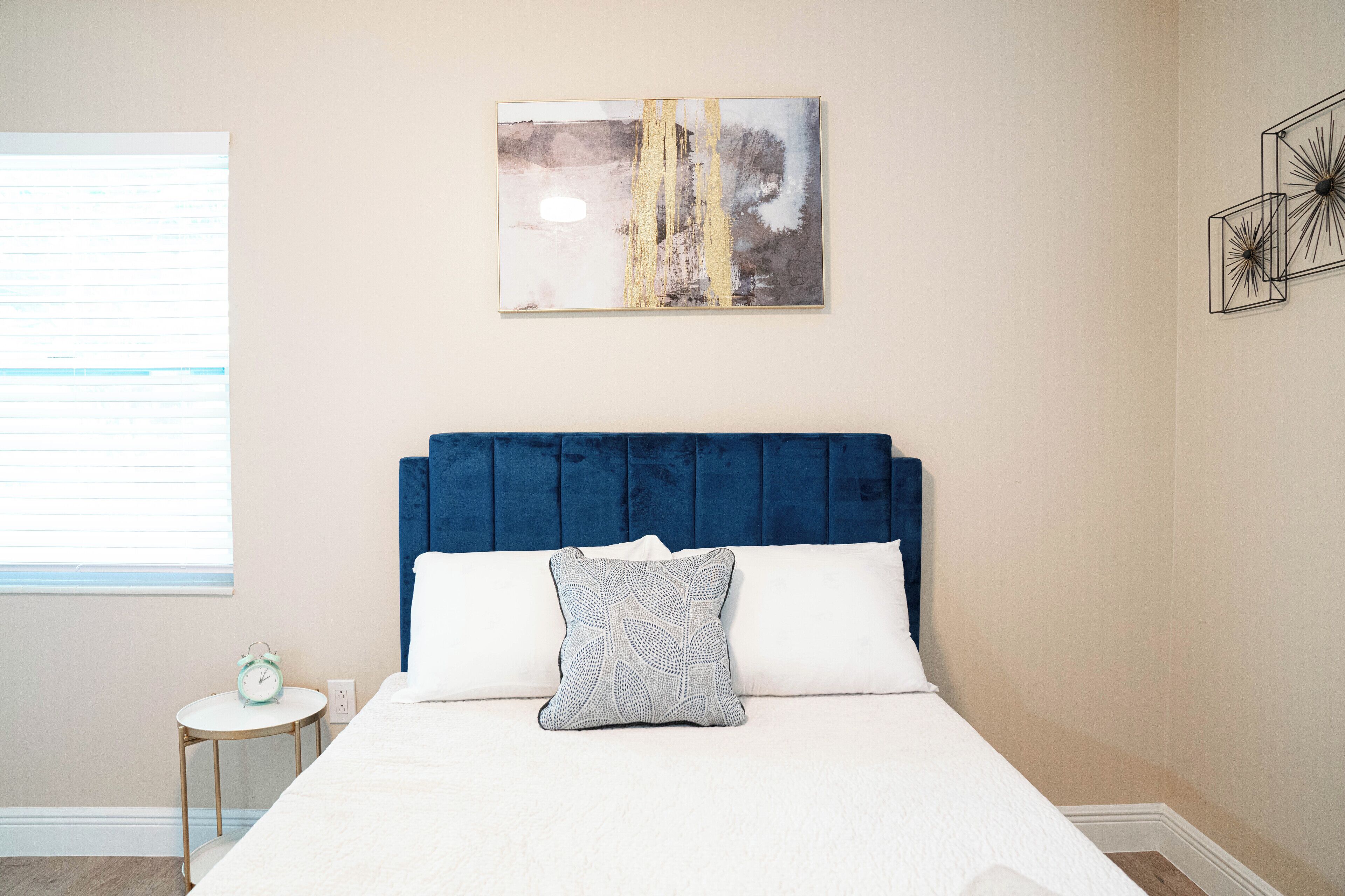 Une chambre à coucher avec un lit, une tête de lit bleue, un oreiller blanc, une petite table ronde avec une horloge, et un tableau abstrait accroché au mur.
