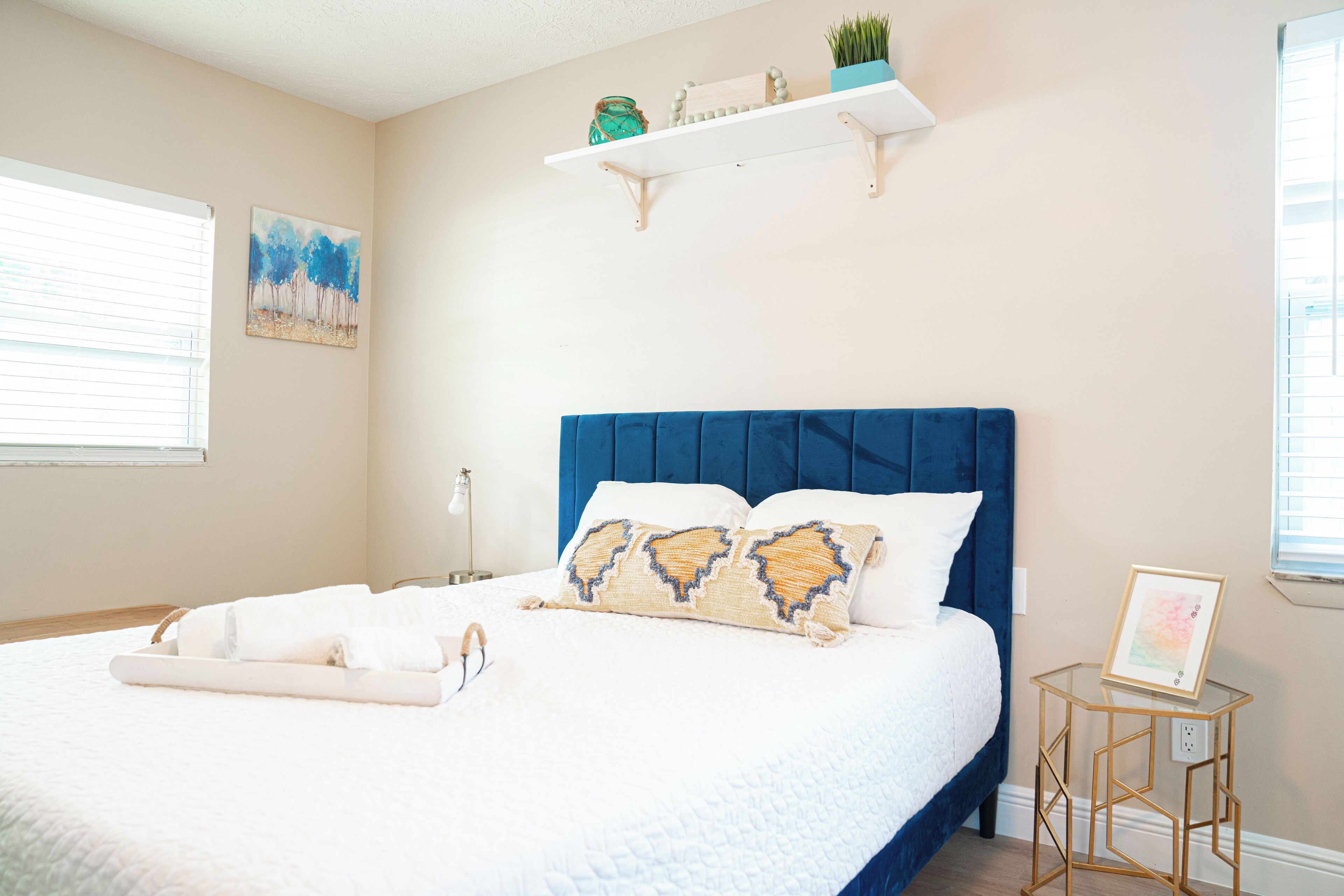 A bedroom with a large bed, a blue headboard, a bedside table with a picture frame, and a wall-mounted shelf with decorative items.