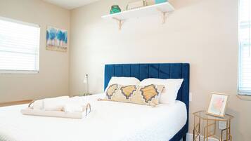 A bedroom with a large bed, a blue headboard, a bedside table with a picture frame, and a wall-mounted shelf with decorative items.