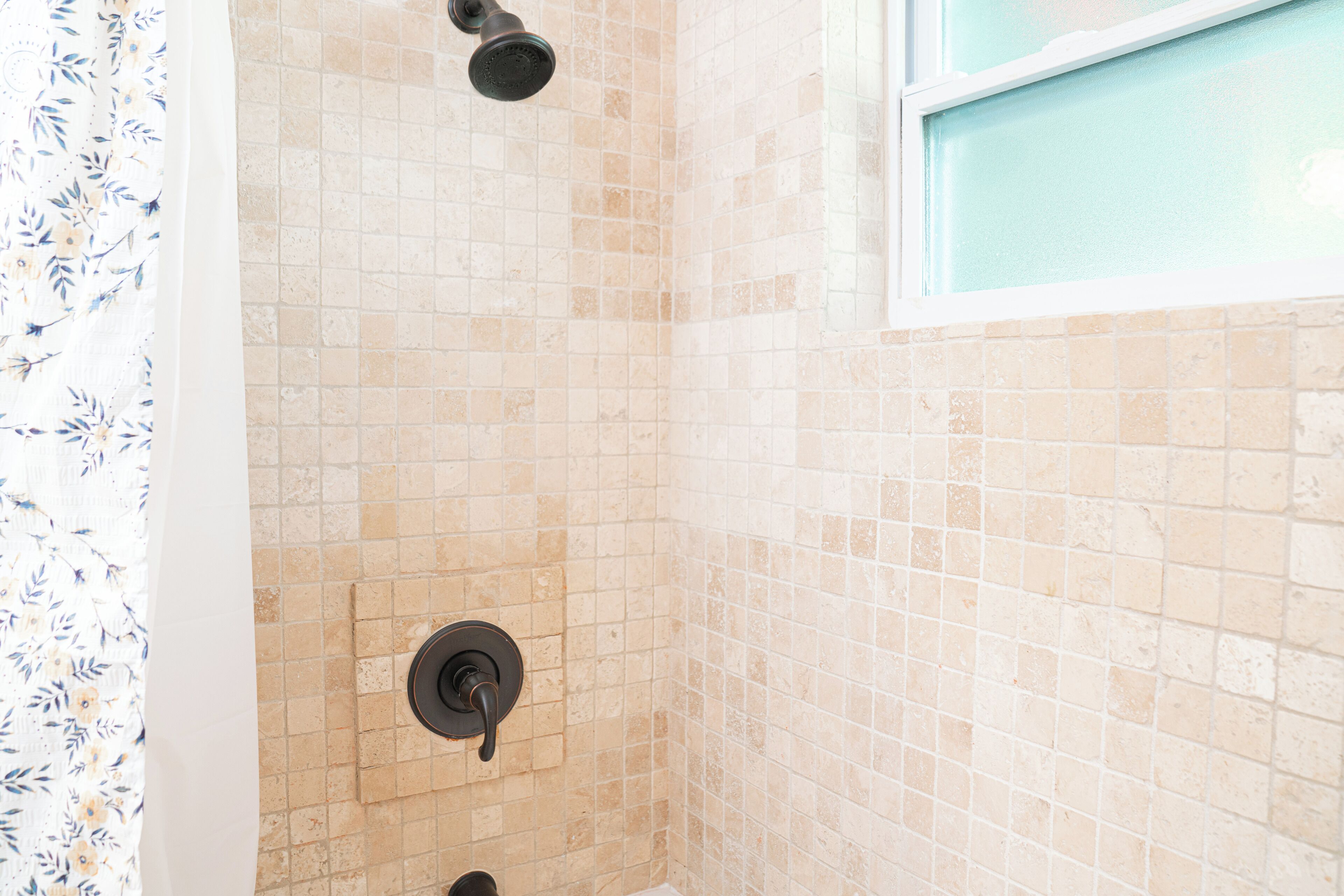 Une salle de bain avec des murs recouverts de carreaux beiges, une pomme de douche et une fenêtre.