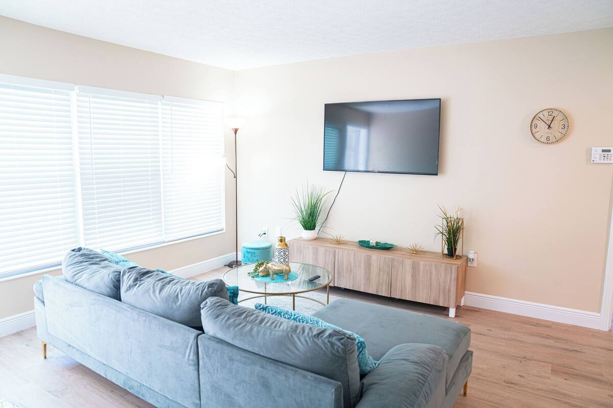 Ein Wohnzimmer mit grauem Sofa, einem runden Couchtisch, einem an der Wand befestigten Fernseher und einer Uhr.