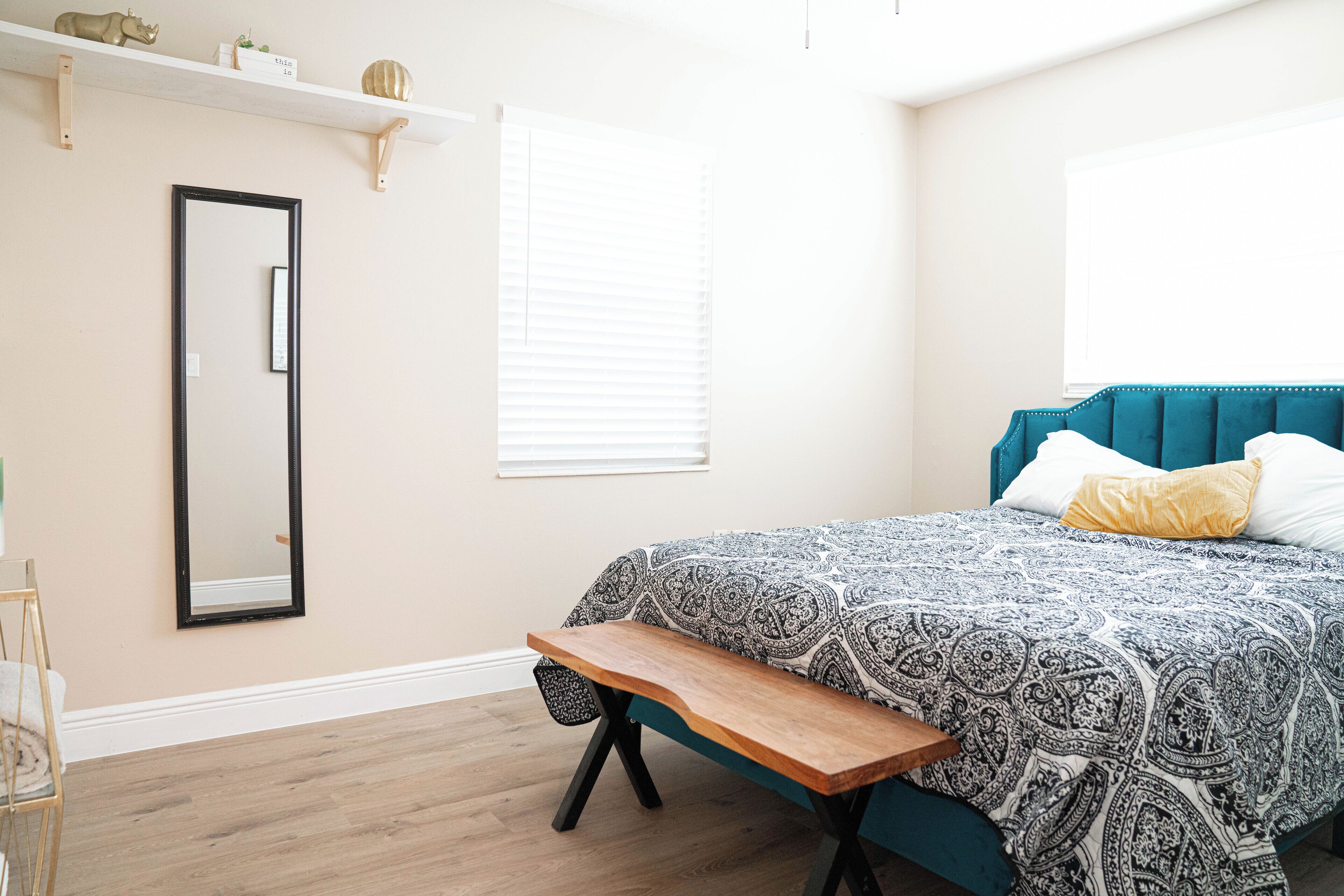 Quarto com cama, um banquinho, espelho e prateleira com objetos decorativos.