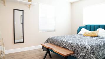 Quarto com cama, um banquinho, espelho e prateleira com objetos decorativos.