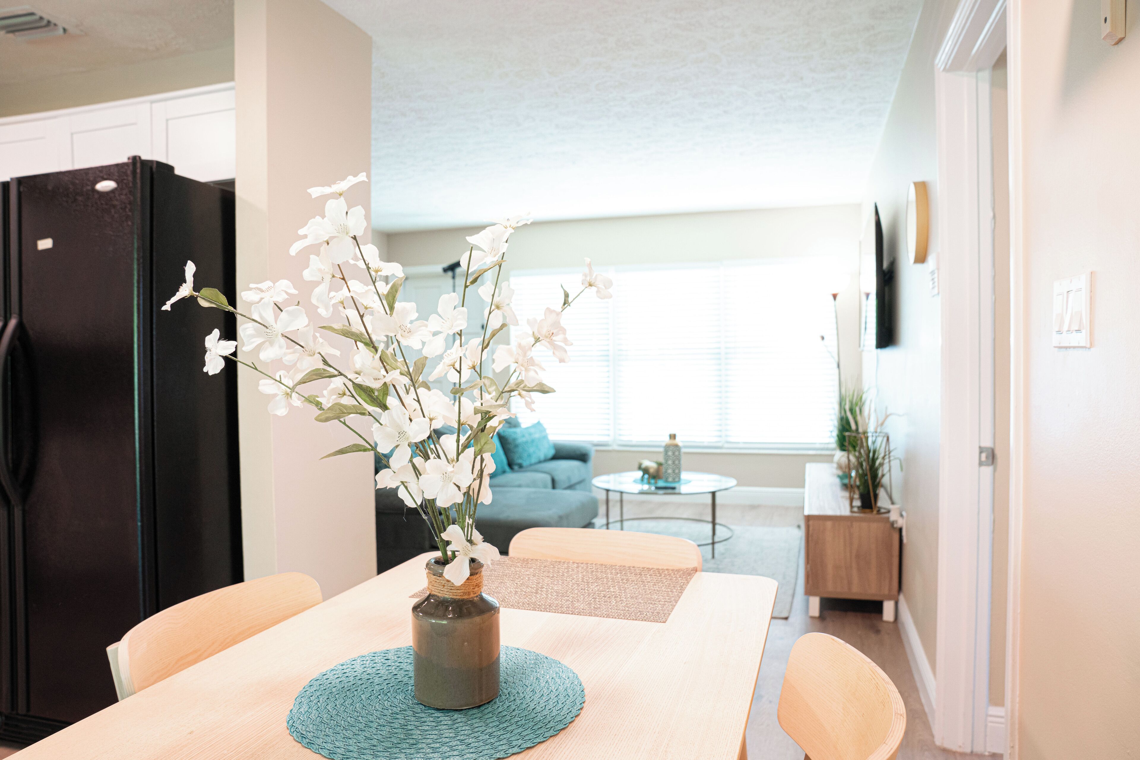 A modern kitchen with a black refrigerator, a dining table with a vase of flowers, and a living area with a sofa and a coffee table.