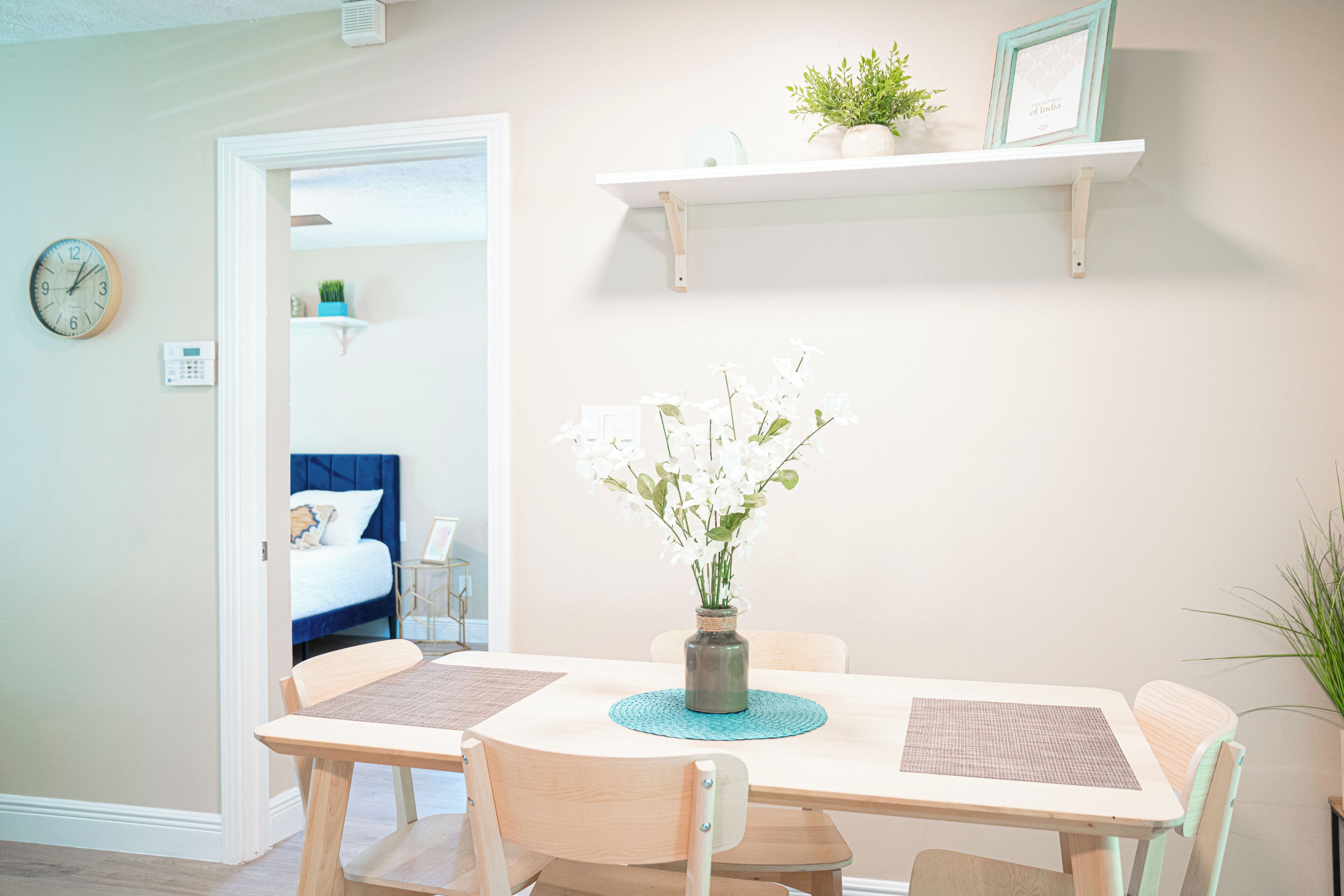 A dining area with a table, chairs, a vase with flowers, a clock, and a bed in the background.