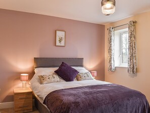 A bedroom with a bed, a nightstand, a lamp, a framed picture on the wall, and a window with curtains.