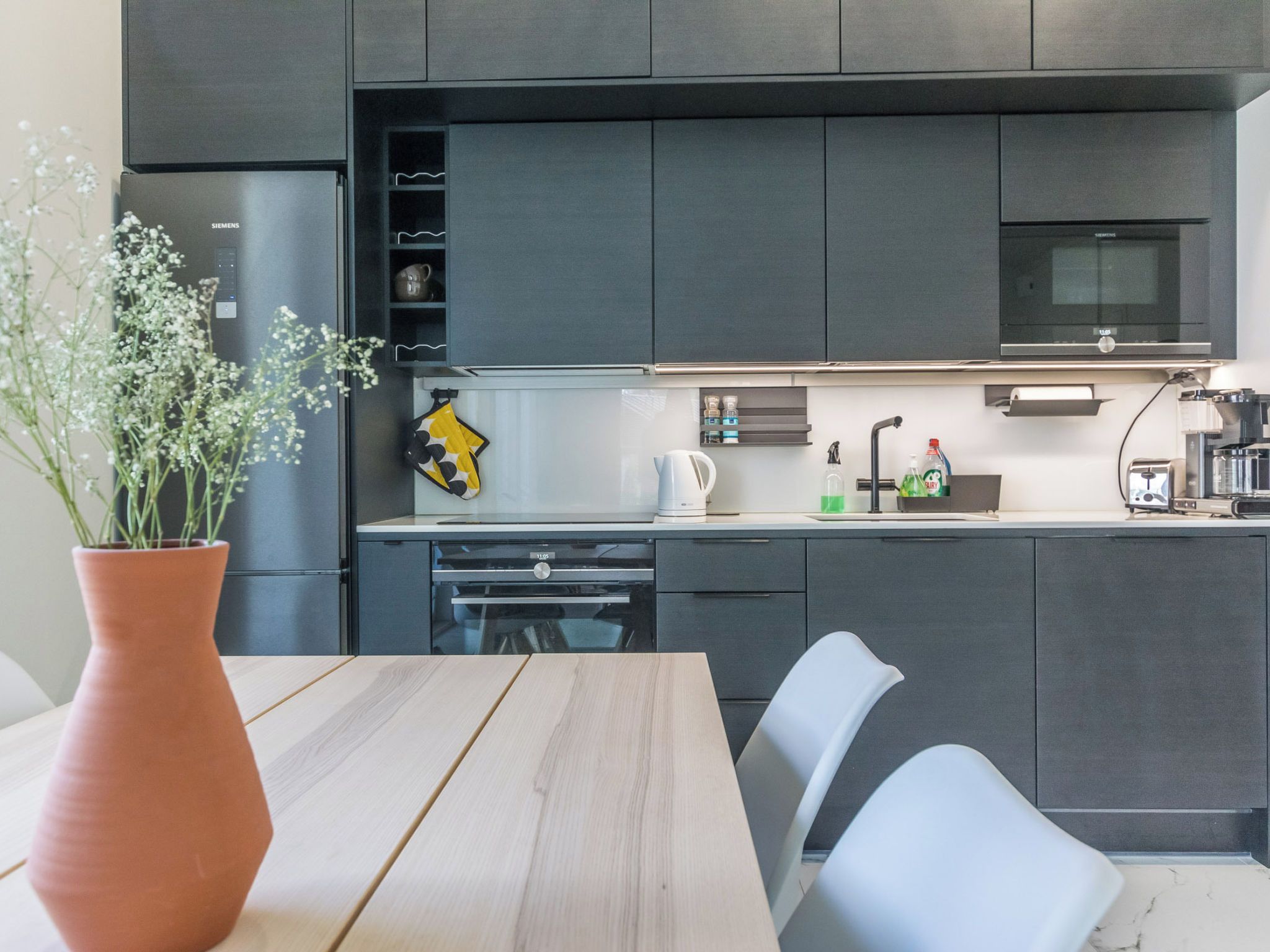 A modern kitchen with dark cabinetry, a stainless steel refrigerator, a microwave, and a dining area with a wooden table and chairs.