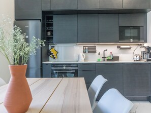 A modern kitchen with dark cabinetry, a stainless steel refrigerator, a microwave, and a dining area with a wooden table and chairs.
