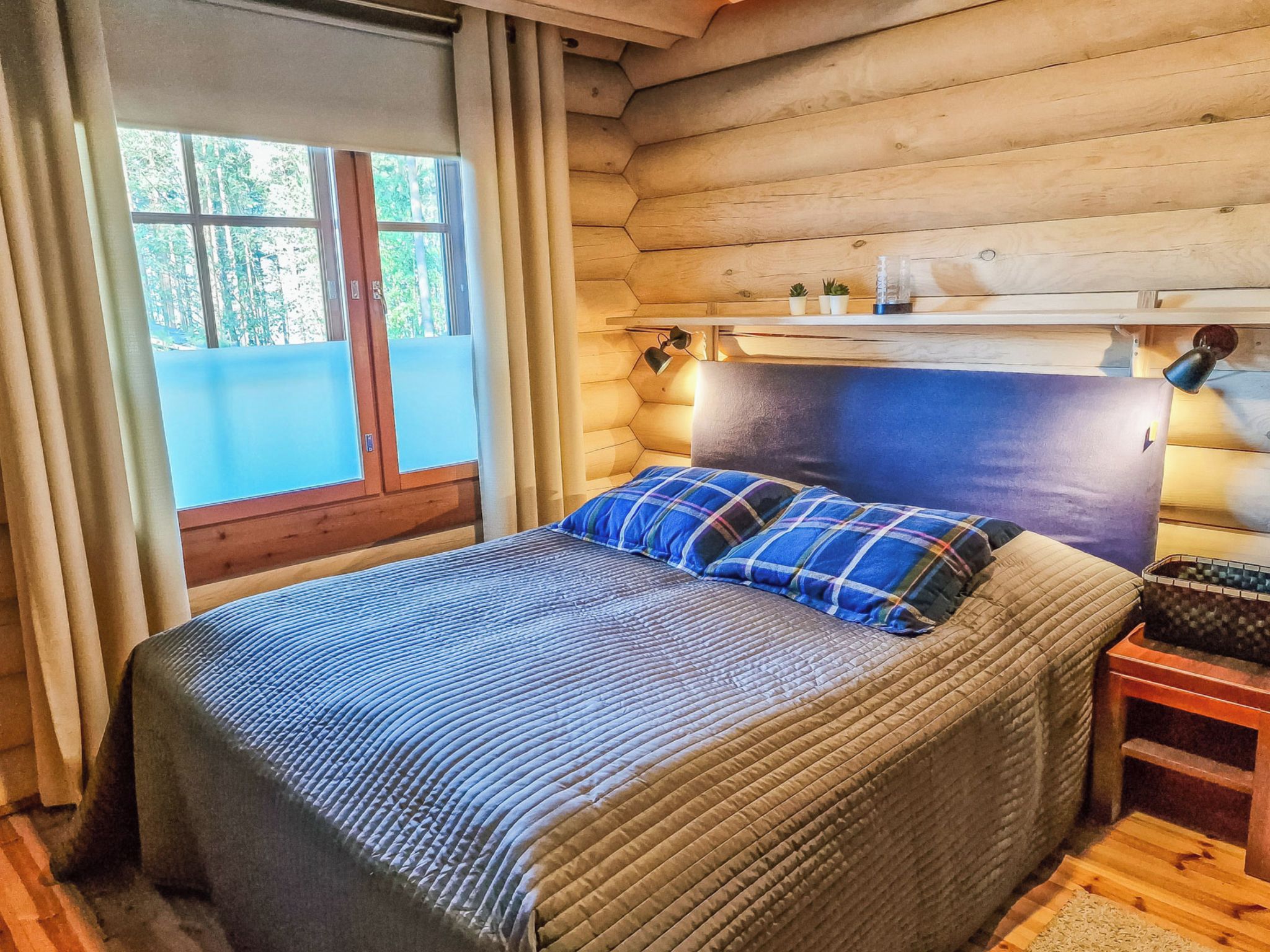 Ein Schlafzimmer in einer Blockhütte mit einem Bett, einem Nachttisch, einem Kopfteil und einem Fenster mit Vorhängen.