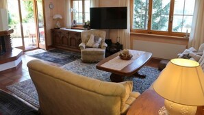 Una sala de estar con sofá, sillón, mesa de centro y televisión. Hay una chimenea, un candelabro y ventanas grandes con cortinas.