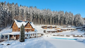 A snow-covered lodge with multiple wooden cabins, a pool, and a forest backdrop.
