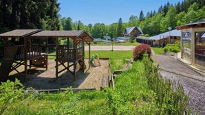 A wooden play structure, a small house, and a glass greenhouse in a lush green setting.