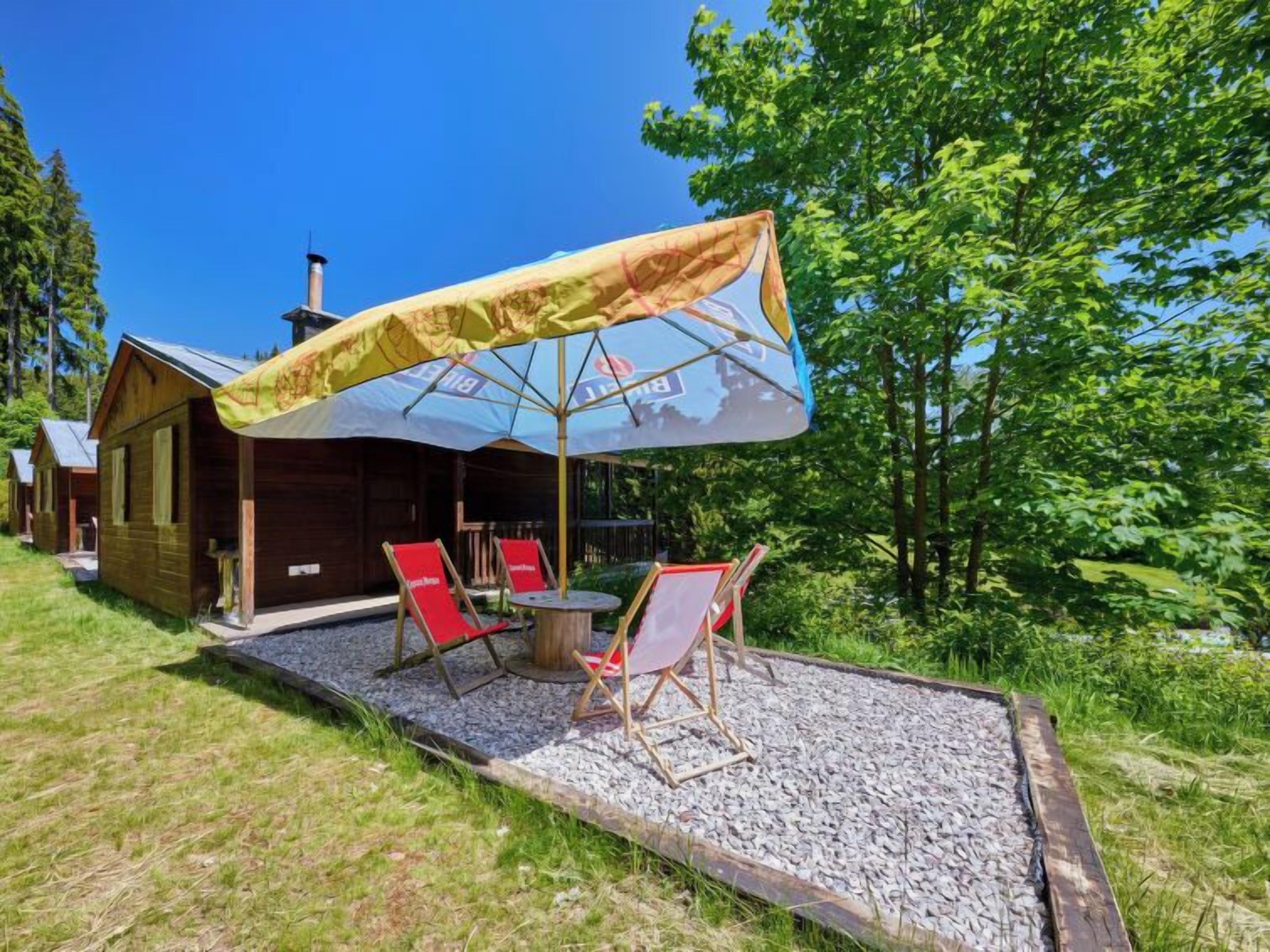 A wooden cabin with a patio area featuring two red lounge chairs and a table, covered by a large umbrella.