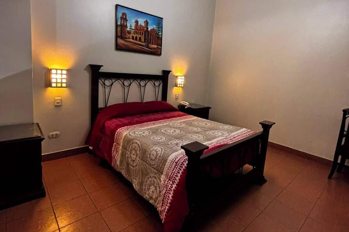 A bedroom with a bed, two bedside lamps, a painting on the wall, and a hanging light fixture.