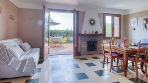 A cozy room with a sofa, a dining table with chairs, a fireplace, and a view through an open door to an outdoor area. - Les Vagues by Interhome (Saint Cyr sur mer Les Lecques)