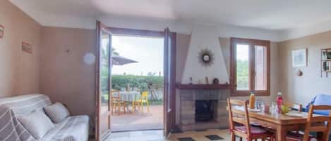 A cozy room with a sofa, a dining table with chairs, a fireplace, and a view through an open door to an outdoor area.