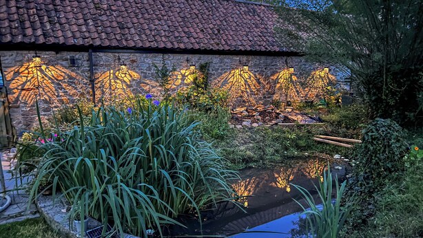 Een gebouw van steen met een pannendak, een vijver en beplanting.