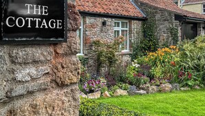 Een stenen vakantiehuisje met een bordje met de tekst 'THE COTTAGE', omringd door een verzorgde tuin met diverse bloemen en een stenen muur.