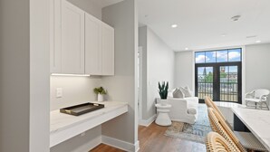 Una cucina moderna con mobili bianchi, un piano di lavoro e una pianta. C'è un'area pranzo con tavolo e sedie bianche e vista su un edificio attraverso ampie finestre.