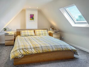 Une chambre avec un plafond en pente, un puits de lumière, un lit recouvert d’une couette à carreaux, des tables de chevet avec des lampes et un tableau accroché au mur.