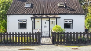 Une petite maison blanche avec un toit noir, une porte d’entrée et une clôture en piquets.