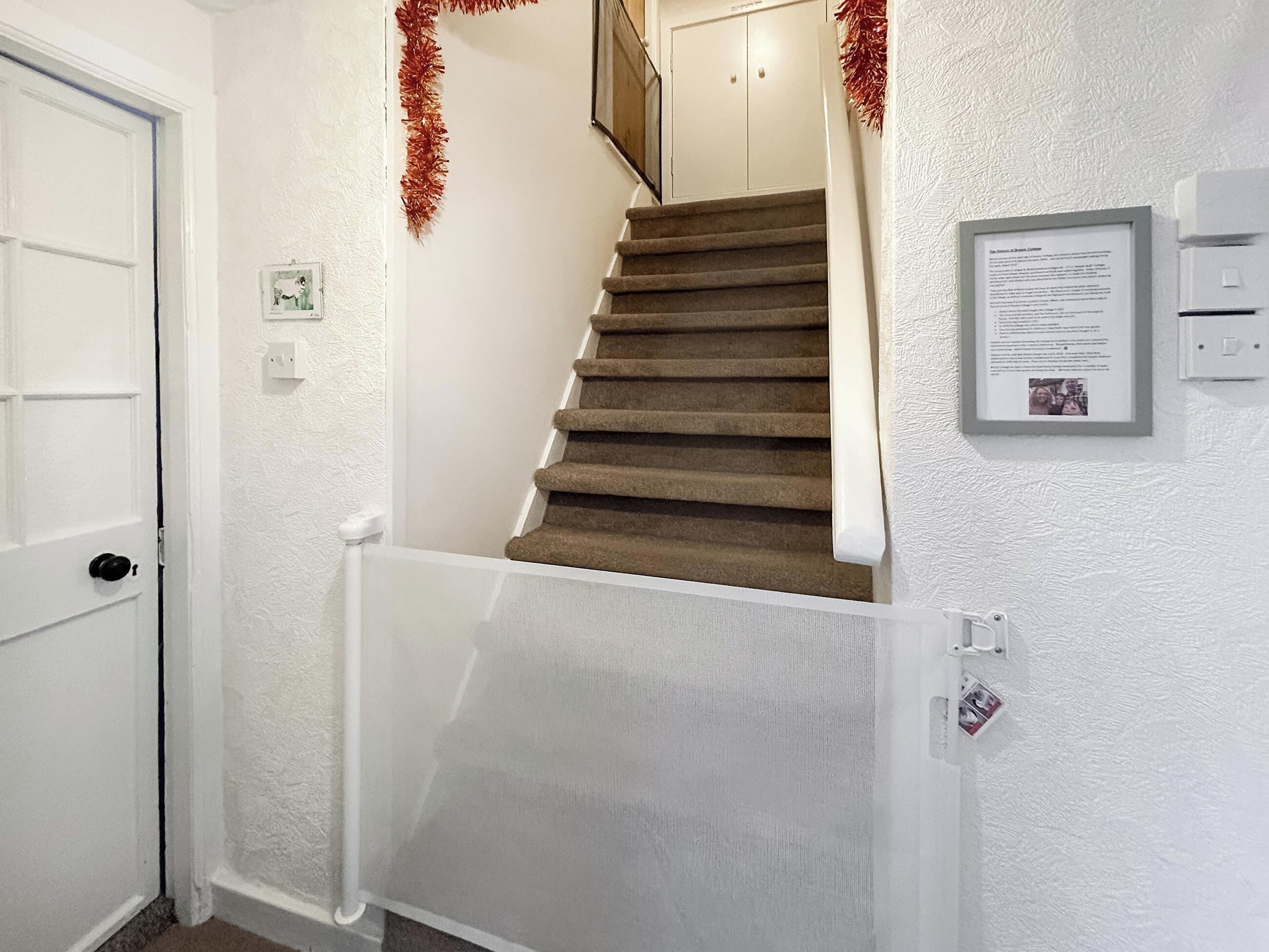 A staircase with a white door on the left, a white wall with a framed notice, and a red tinsel decoration.