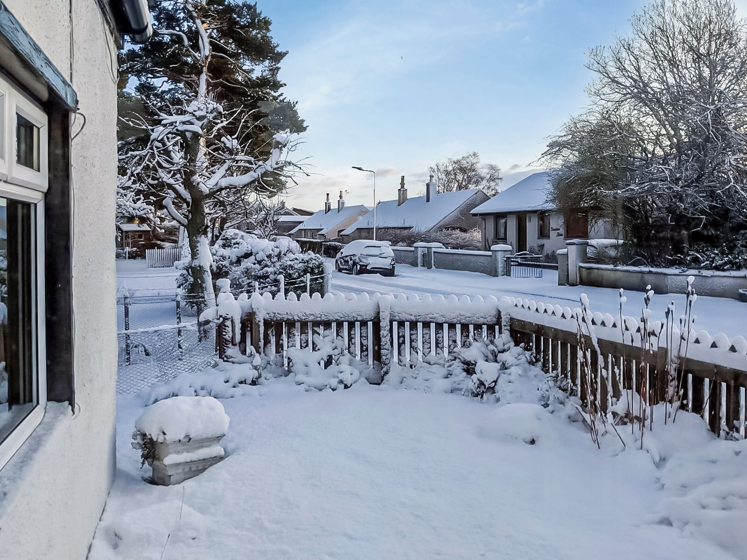 A residential street with snow-covered houses and trees, a wooden fence, and a parked car.