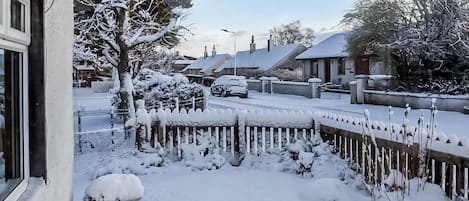 Calle residencial con casas y árboles cubiertos de nieve, una valla de madera y un coche estacionado.