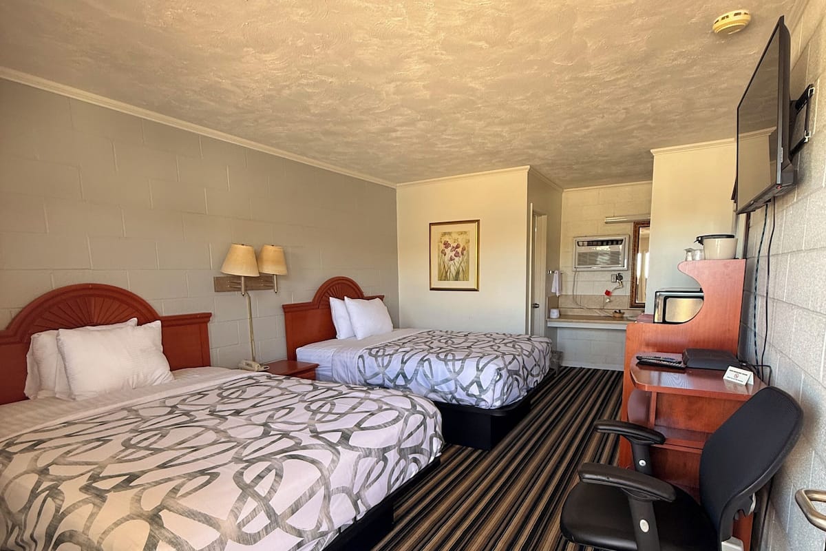 A hotel room with two beds, a desk, a chair, a lamp, and a television.