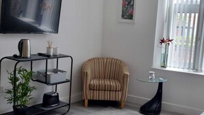 A living room with a striped armchair, a small round table, a TV mounted on the wall, and a window with blinds. - Charming 1-bed Studio in Preston (Preston)