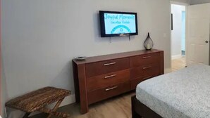 A bedroom with a bed, a wooden dresser, a small table, and a TV on the wall displaying 'Joyful Moments'.