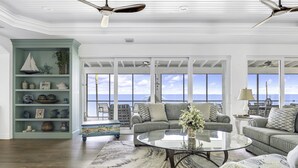 A spacious living room with a large window overlooking the sea, a teal bookshelf, and a round glass coffee table. - Life Changing Views from this Luxury 3 Bedroom Bayfront Home! (Captiva)