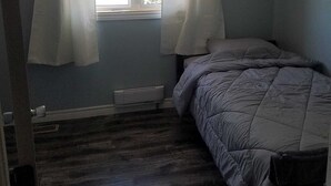 Une petite pièce, peu meublée, avec un lit simple, une fenêtre avec des rideaux et un radiateur au sol.