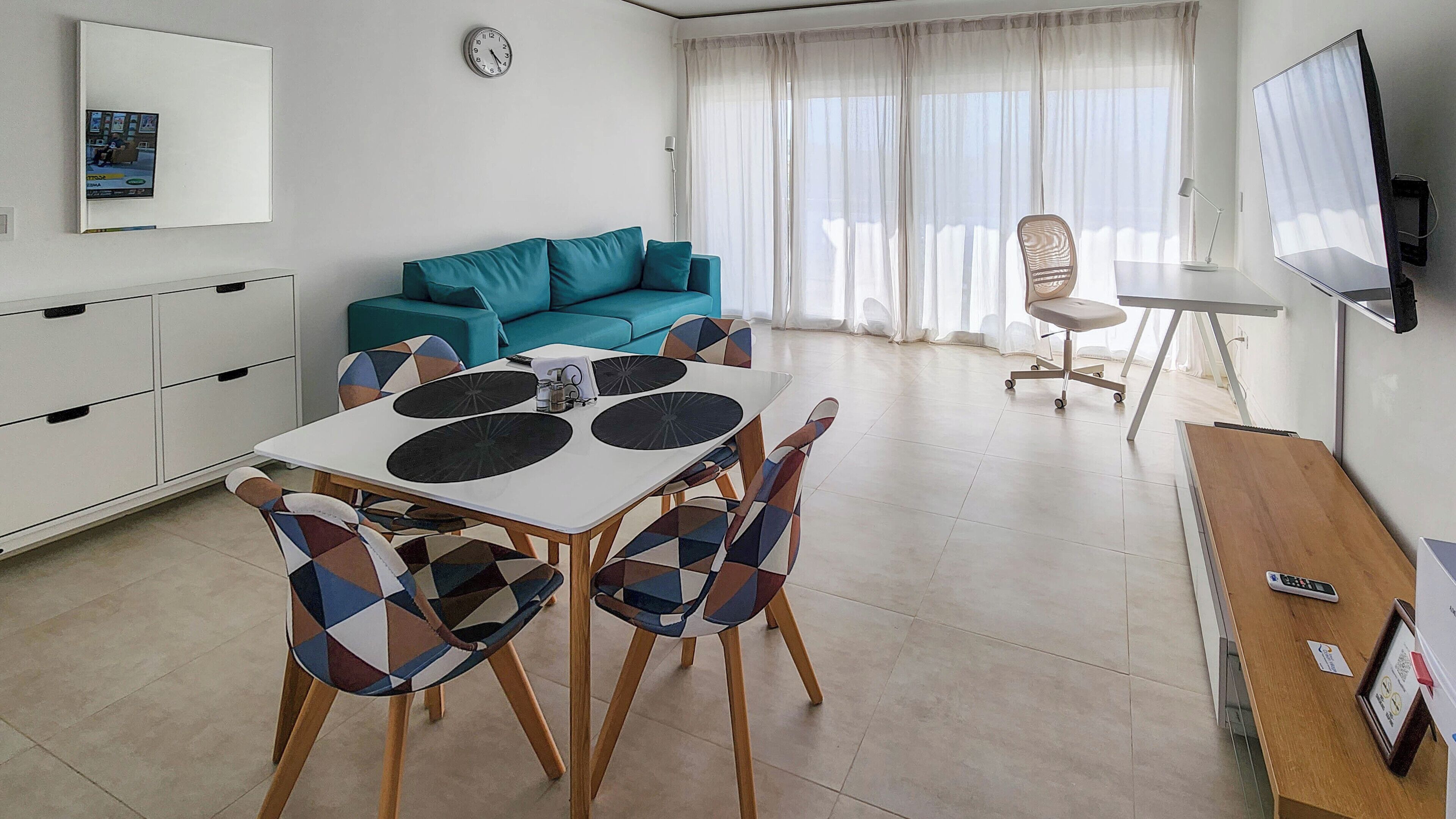 Un salon moderne avec un canapé de couleur turquoise, une table à manger entourée de chaises à motifs, un téléviseur fixé au mur et une grande fenêtre ornée de rideaux légers.