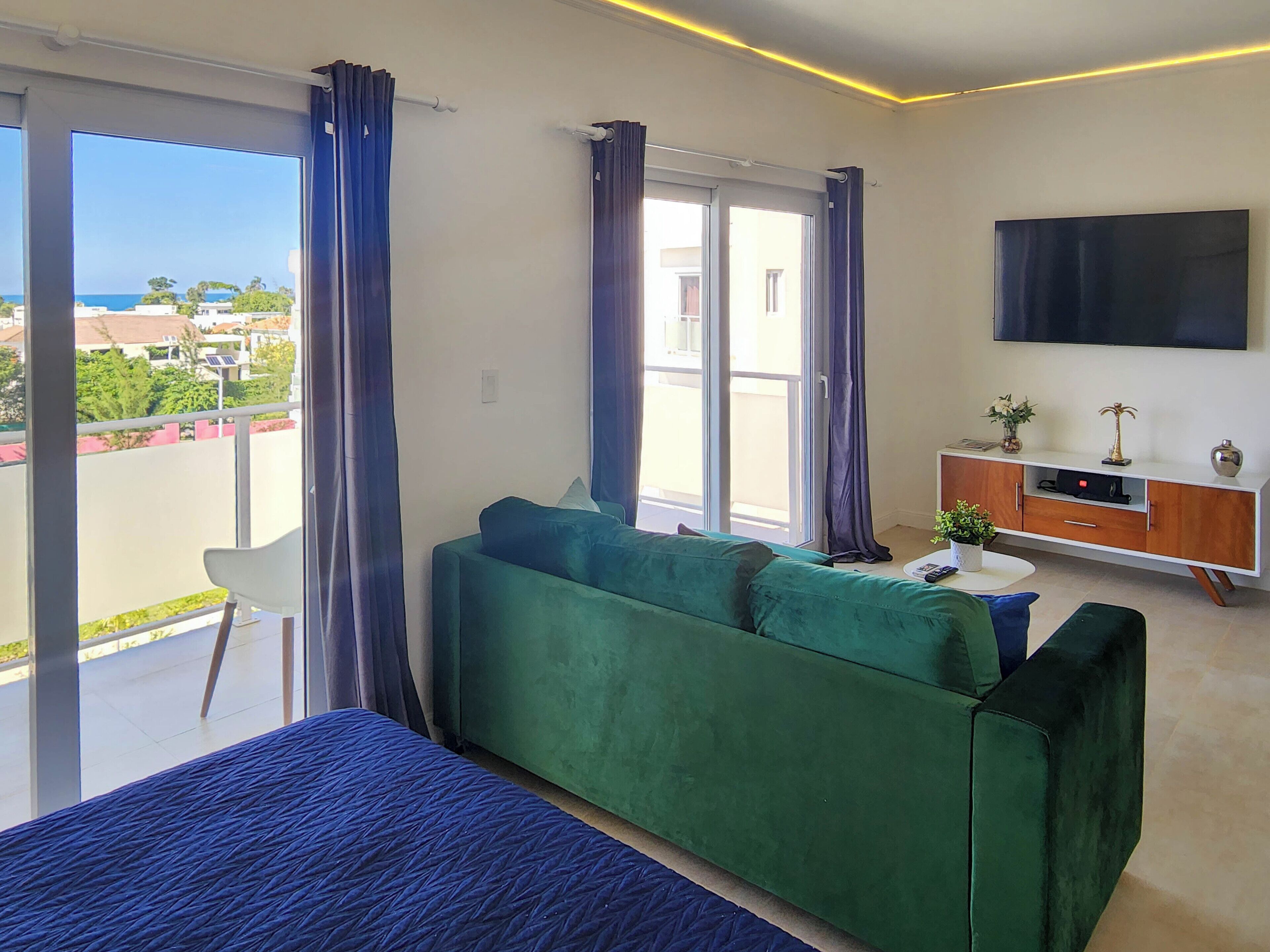 A modern living room with a green sofa, a flat-screen TV, and a balcony with curtains.