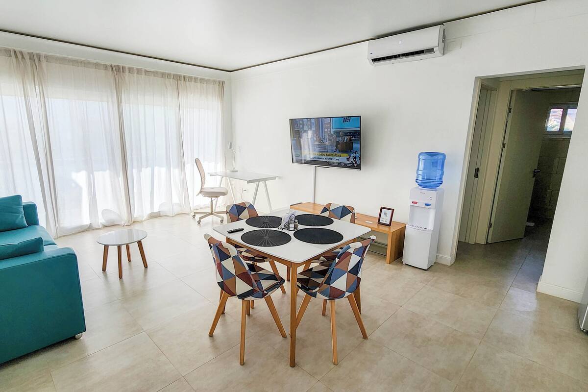 Een moderne woonkamer met een blauwgroene bank, een eettafel met vier stoelen, een aan de muur bevestigde televisie en een waterdispenser.