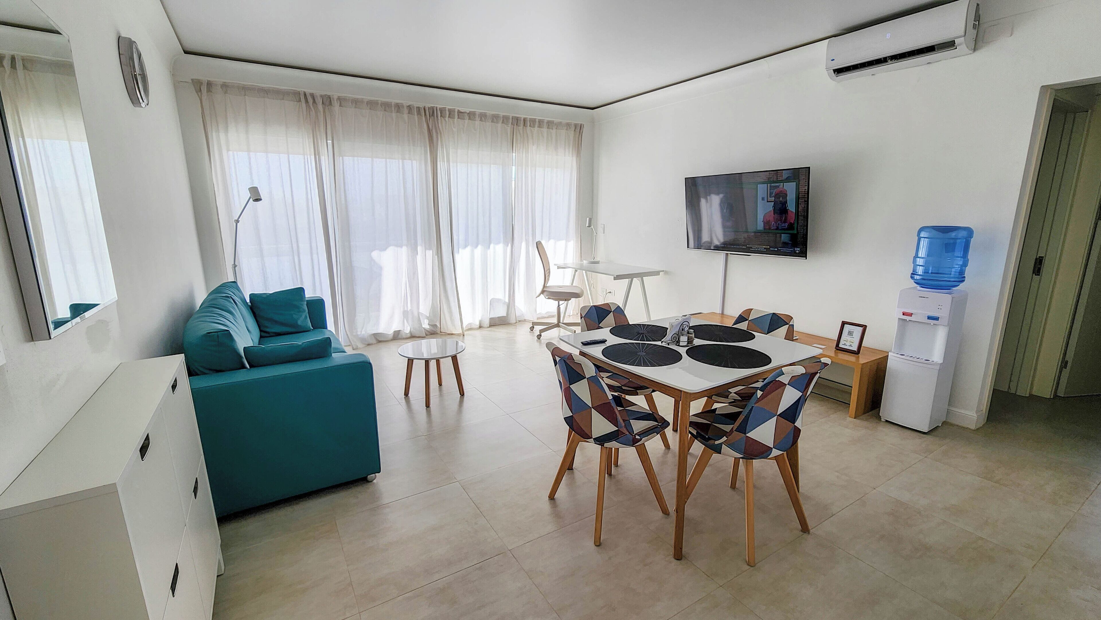 Una sala de estar moderna con un sofá color turquesa, una mesa de comedor con sillas, un televisor de pantalla plana montado en la pared y un dispensador de agua.