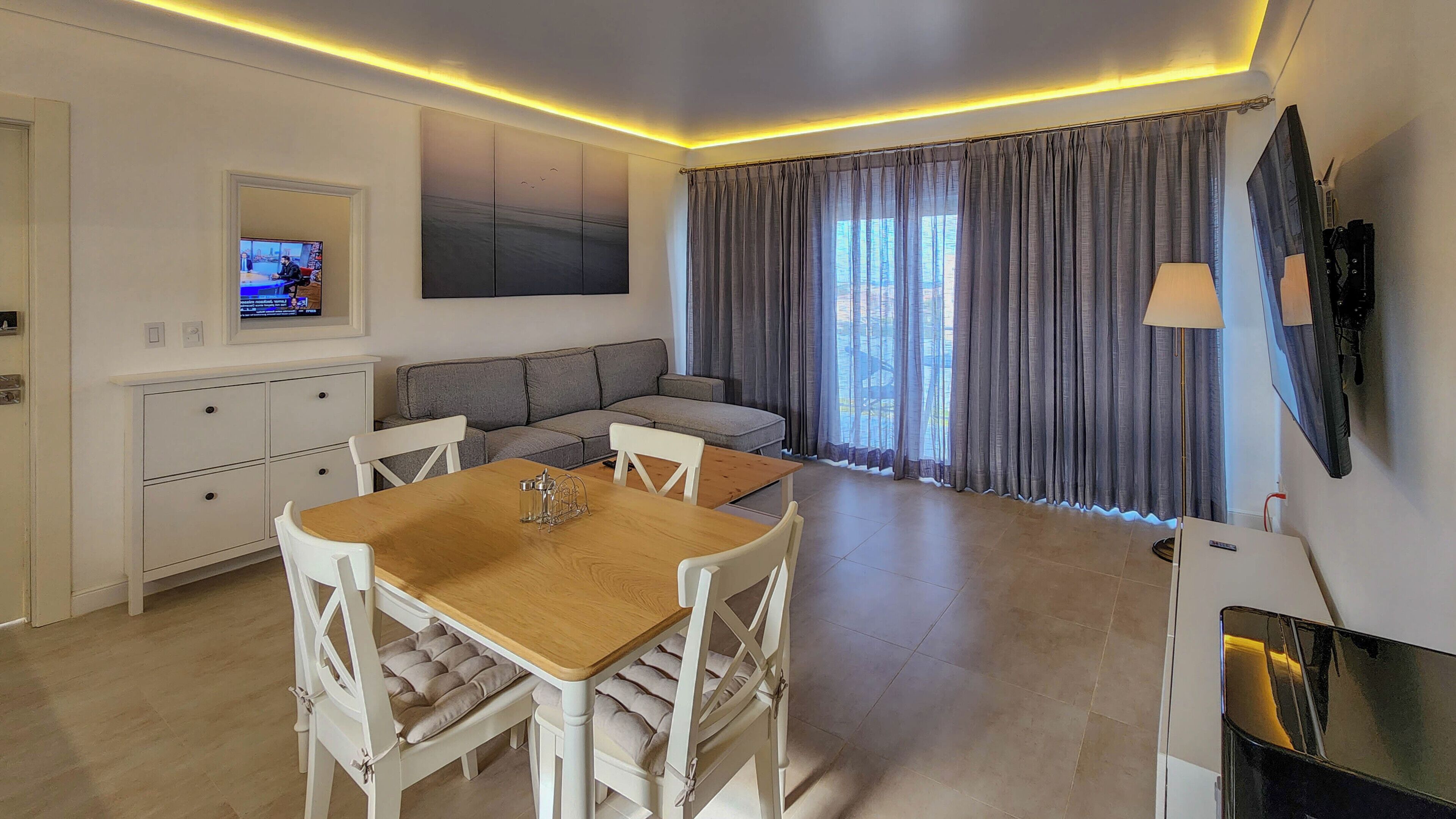 A modern living room with a wooden dining table, white chairs, a grey sofa, and a TV mounted on the wall.