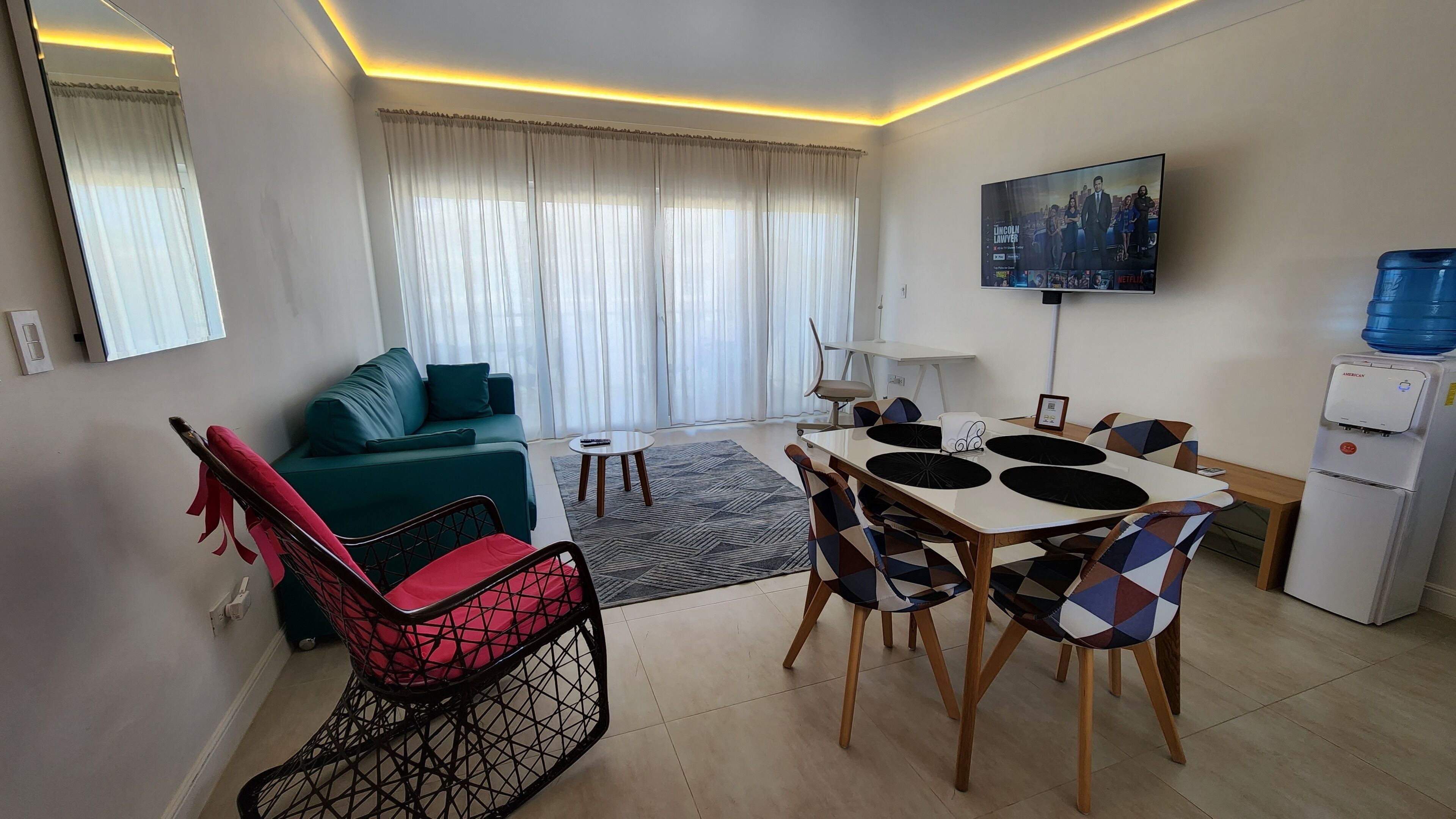 A modern living room with a sofa, a dining table with chairs, a TV mounted on the wall, and a water dispenser.